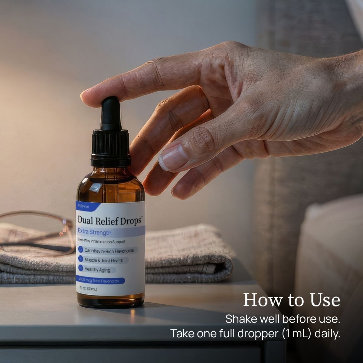 A hand reaches toward a bottle of Dual Relief™ Drops on a tabletop, with folded fabric and eyeglasses in the background and overlaid text reading 'How to Use' and 'Shake well before use. Take one full dropper (1 mL) daily.'